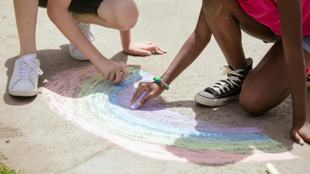 Mini Masterminds Child Care Two individuals are drawing a rainbow with colored chalk on a concrete surface, showcasing the imaginative spirit fostered by Youth Programs and Mini Masterminds.