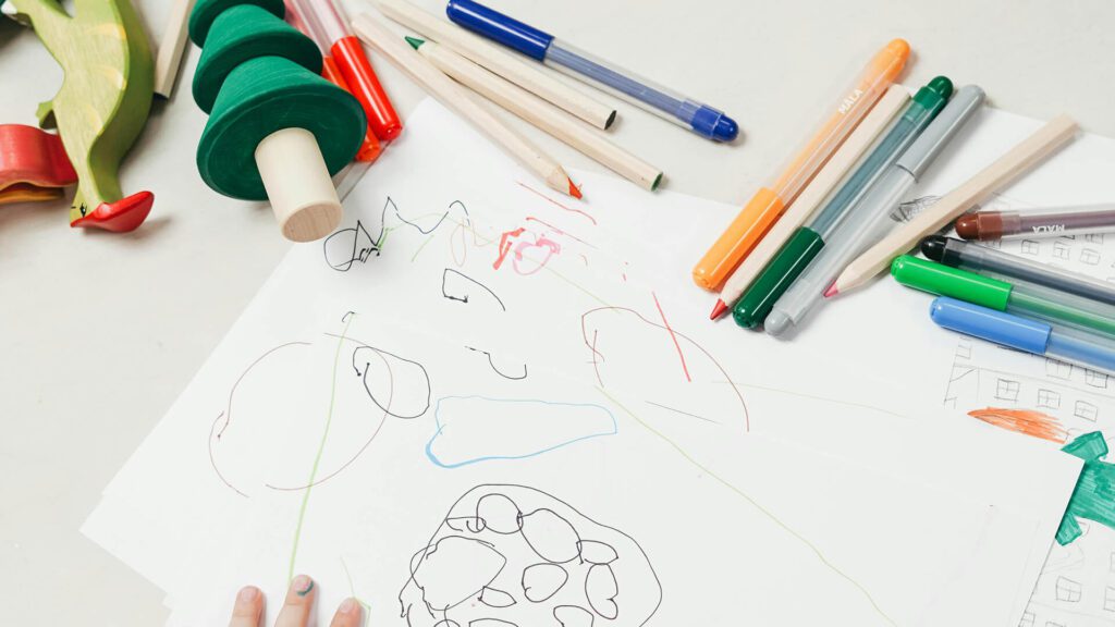 Mini Masterminds Child Care A child's drawing on paper, surrounded by various colored markers, pens, and wooden toys on a white table, showcases the creativity encouraged in our Youth Programs.