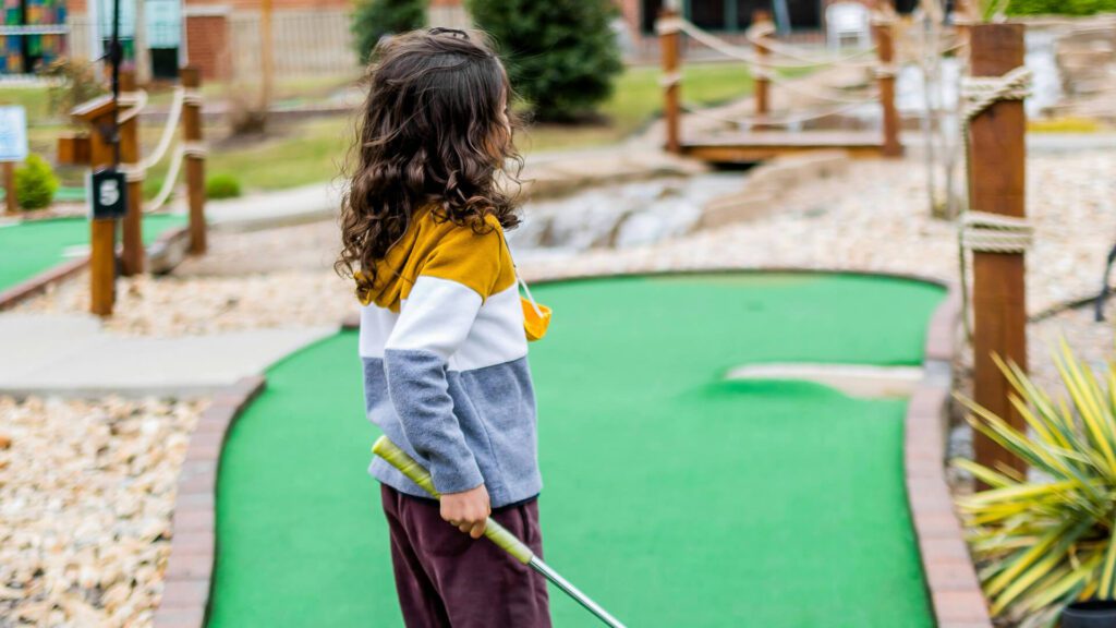 Mini Masterminds Child Care A Child with long curly hair in a color-block hoodie, a potential Mini Mastermind in the making, holds a mini-golf putter while standing on a mini-golf course, facing away from the camera.