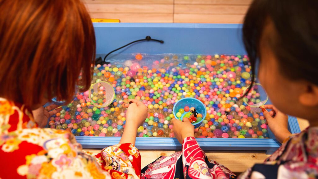 Mini Masterminds Child Care Two children, dressed in colorful kimonos, play with small, multicolored balls in a blue water-filled container using scoops. This immersive activity is just one of many engaging experiences offered by our Mini Masterminds youth programs.