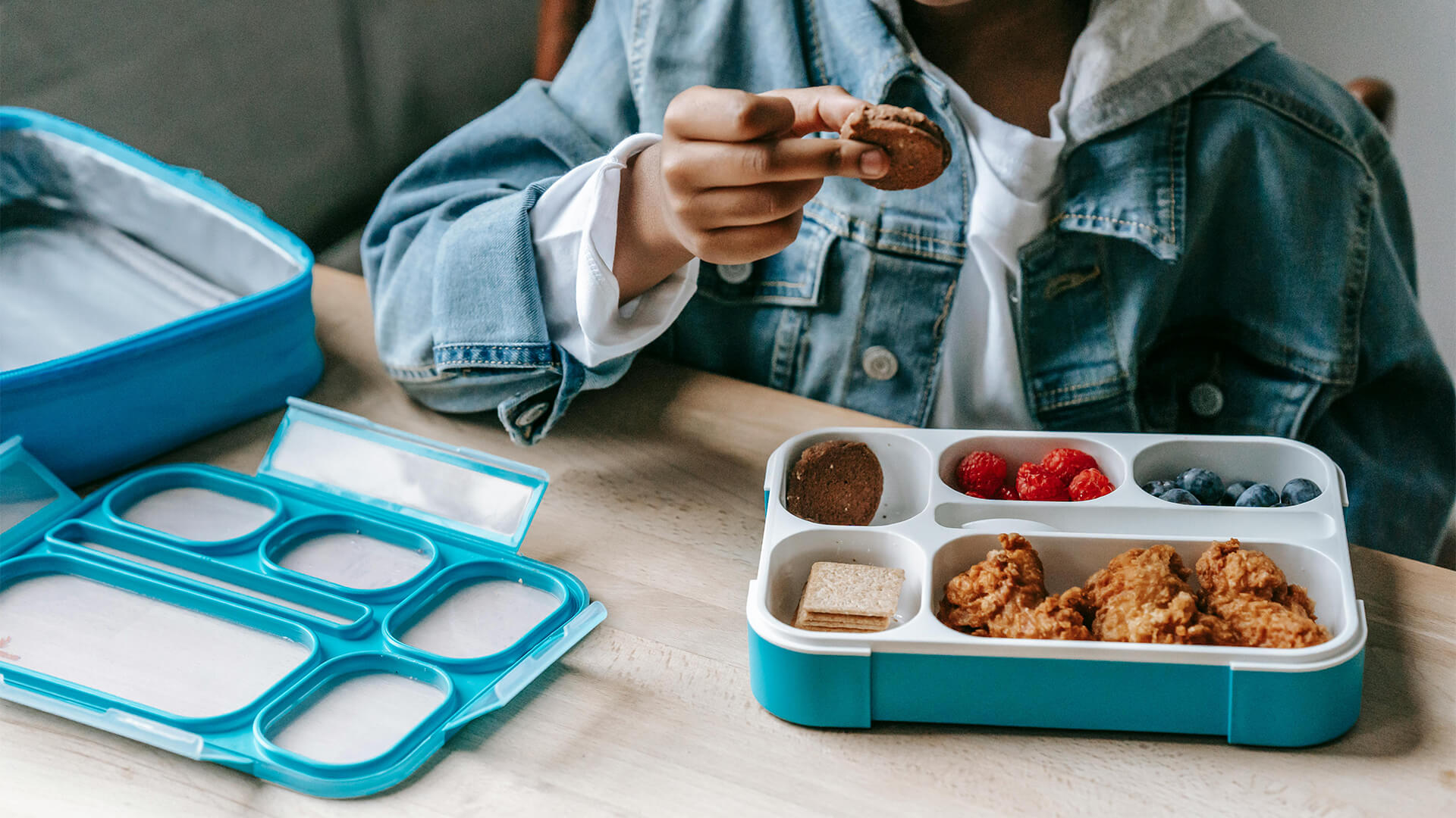 Mini Masterminds Child Care A person in a denim jacket holds a chocolate cookie while sitting at a table with an open lunchbox containing fried chicken, berries, crackers, and cookies. An empty lunch container from Mini Masterminds sits nearby.