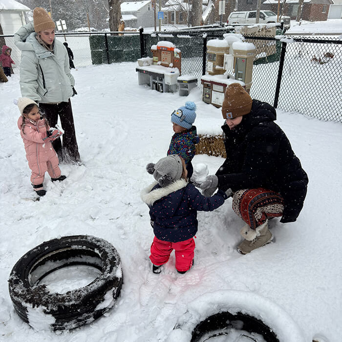Mini Masterminds Child Care Two adults and three young children play outdoors in the snow near some large tires and toy kitchen sets, while snow falls around them.