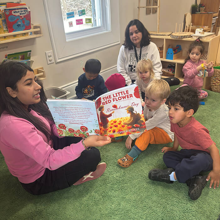 Mini Masterminds Child Care A woman reads "The Little Red Flower" to a group of young children sitting on a green carpet in a classroom, while another adult observes.