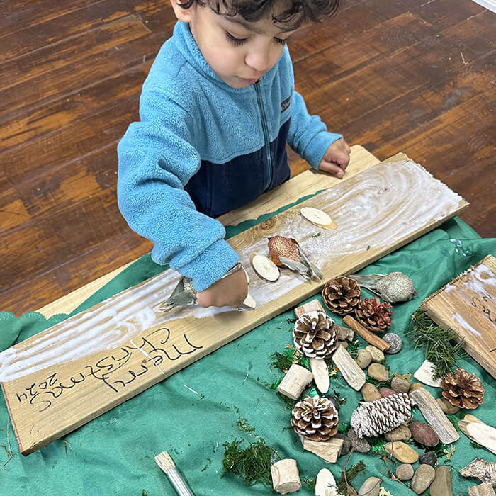 Mini Masterminds Child Care A young child in a blue fleece decorates a wooden plank with natural materials like pine cones, rocks, and sticks on a table. The plank has writing that appears to say "Merry Christmas 2024.