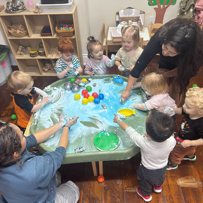 Mini Masterminds Child Care A group of young children and two adults play with colorful balls and sensory materials at a water table in a classroom setting.
