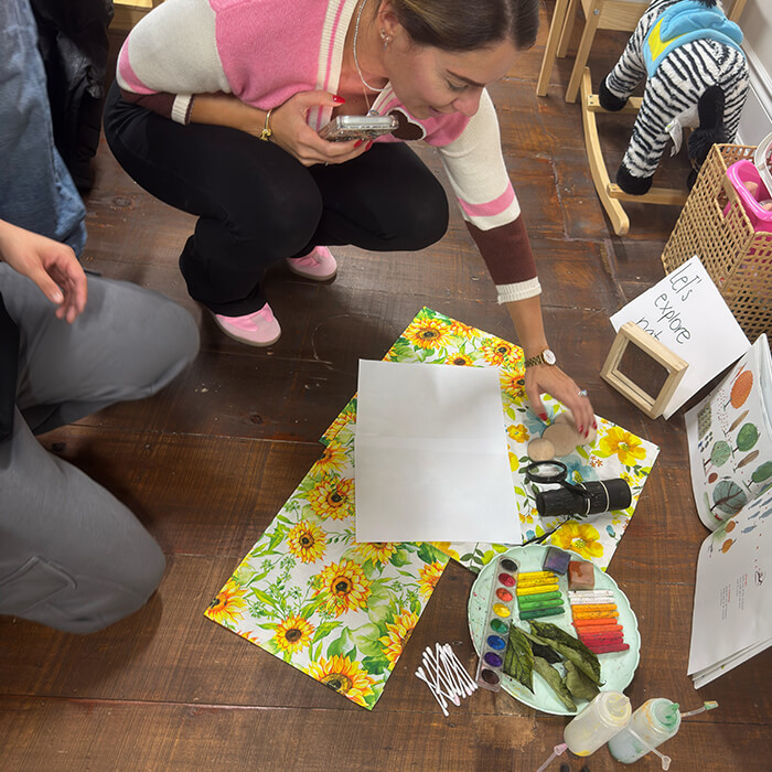 Mini Masterminds Child Care Two people prepare art supplies, including paint, leaves, and paper, on a sunflower-patterned mat on a wooden floor. A sign reads “Ed’s Corner.”.