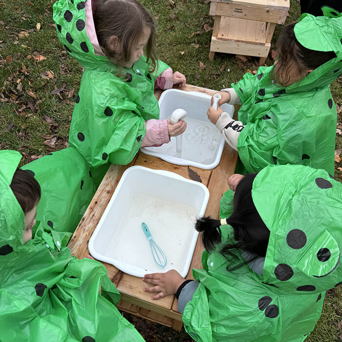 Mini Masterminds Child Care Four children wearing green rain ponchos with black spots play with kitchen utensils in two white tubs of water outdoors.