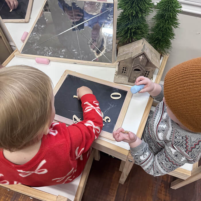 Mini Masterminds Child Care Two young children draw numbers on small chalkboards at a table with a mirror, a model house, and small faux pine trees.