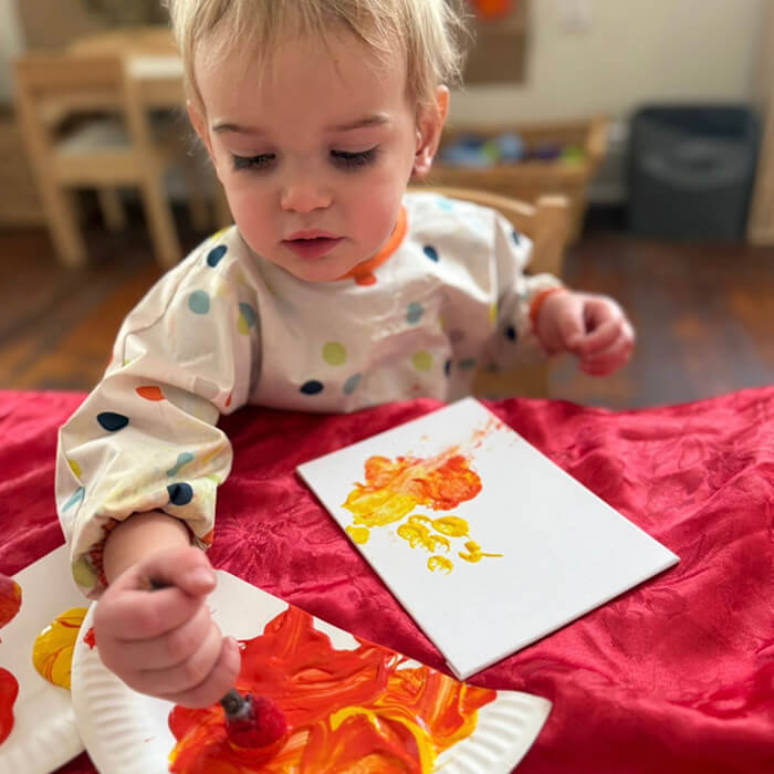 Mini Masterminds Child Care A young child in a polka dot smock paints with red and yellow colors on a piece of paper at a table.
