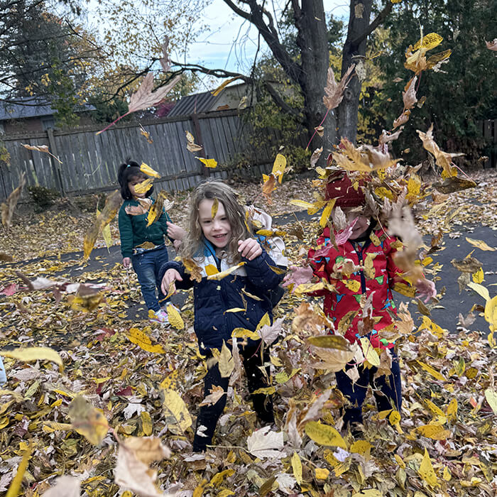 Mini Masterminds Child Care Three children play outside, throwing and surrounded by fallen autumn leaves, with trees and a fence in the background.