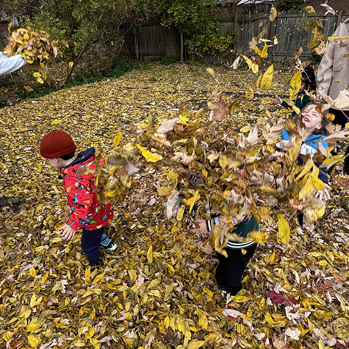 Mini Masterminds Child Care Children play in a yard covered with yellow autumn leaves, tossing leaves in the air and smiling among trees and a wooden fence.