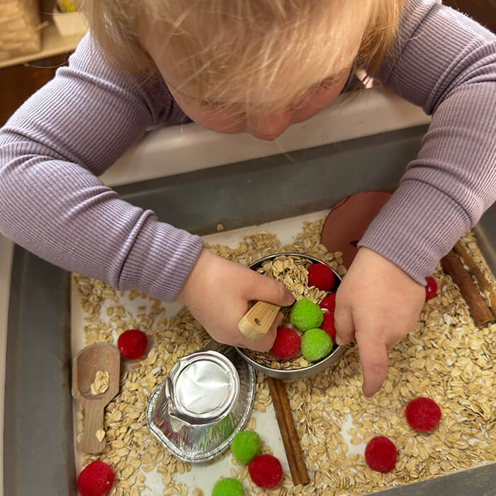 Mini Masterminds Child Care A young child in a purple shirt plays with oats, red and green pom-poms, a wooden scoop, foil cups, and cinnamon sticks in a sensory bin.
