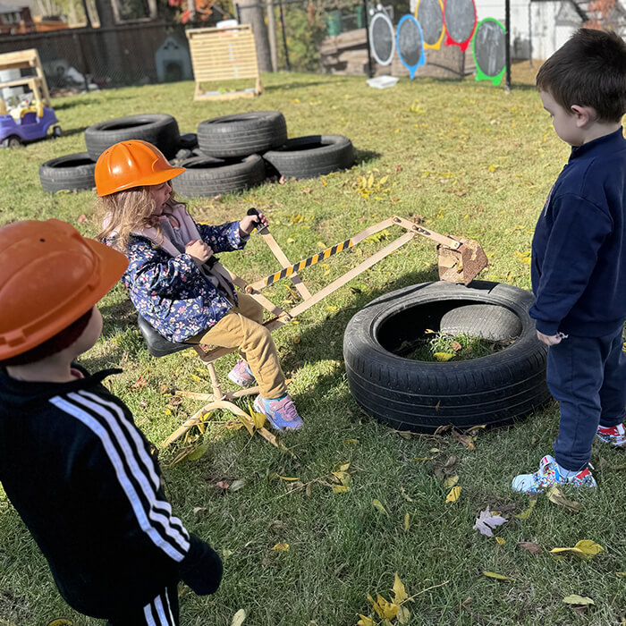 Mini Masterminds Child Care Three children play outside; one girl wearing an orange hard hat operates a toy digger with tires, while two boys watch.
