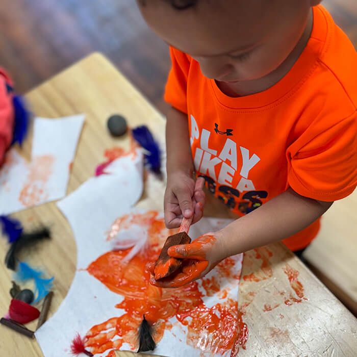 Mini Masterminds Child Care A young child in an orange shirt paints on paper with a brush, surrounded by feathers, rocks, and paint on a wooden table.
