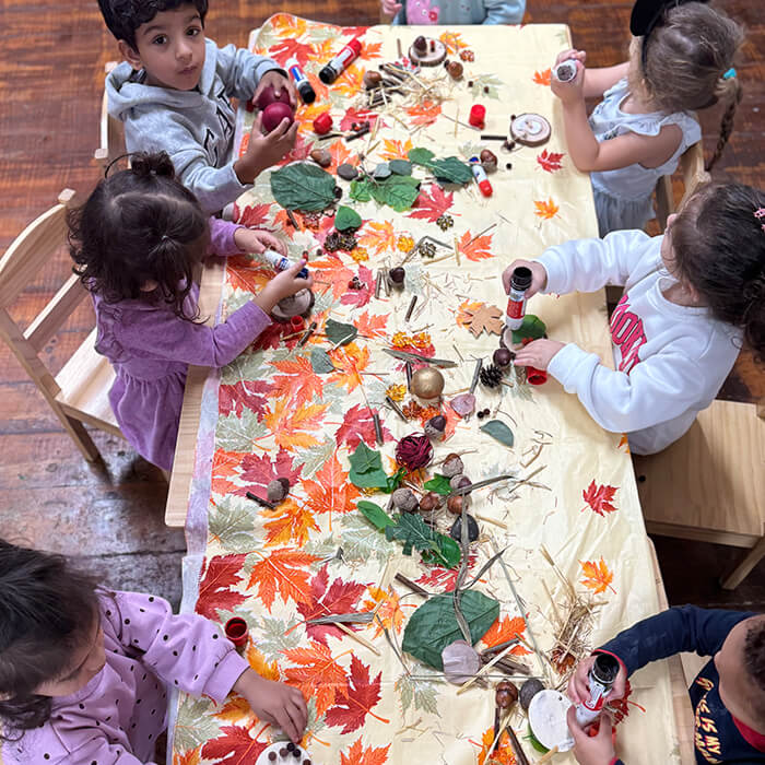 Mini Masterminds Child Care Five young children sit around a table covered with autumn leaves and natural materials, engaging in a craft activity with paint and glue.