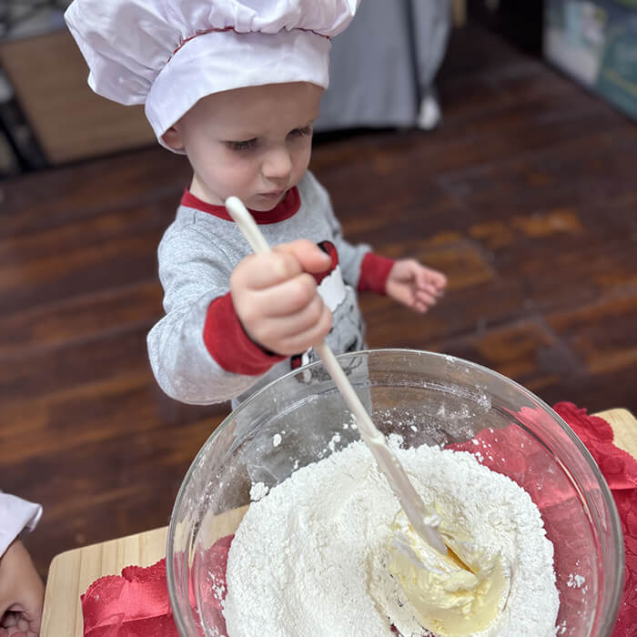 Mini Masterminds Child Care A young child wearing a chef’s hat stirs flour and butter in a large glass bowl with a wooden spoon on a wooden table.