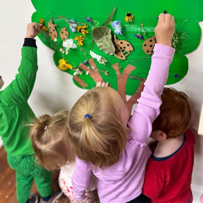 Mini Masterminds Child Care Three young children are decorating a green paper tree on a wall with leaves, flowers, and other craft materials.