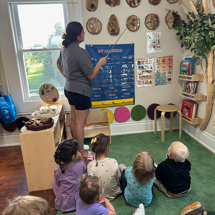Mini Masterminds Child Care A teacher stands and points to a calendar chart on the wall while a group of young children sit on the floor and watch attentively in a classroom.