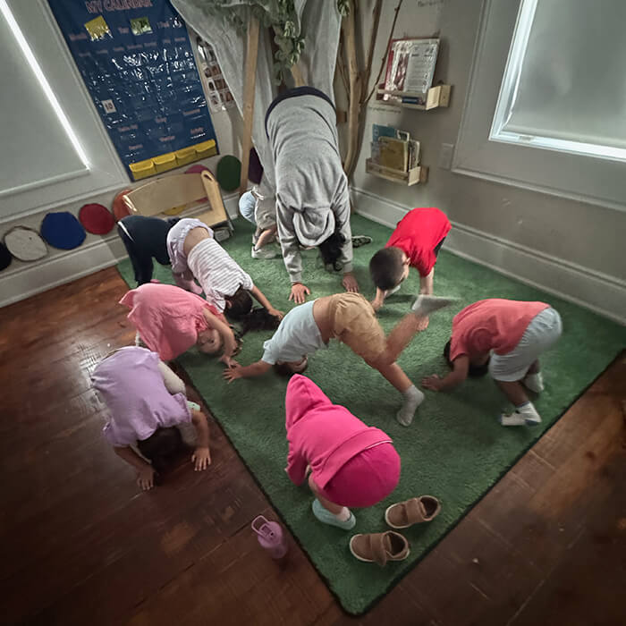 Mini Masterminds Child Care A group of young children and an adult practice yoga on a green rug in a classroom, all bending forward with hands and feet on the floor.