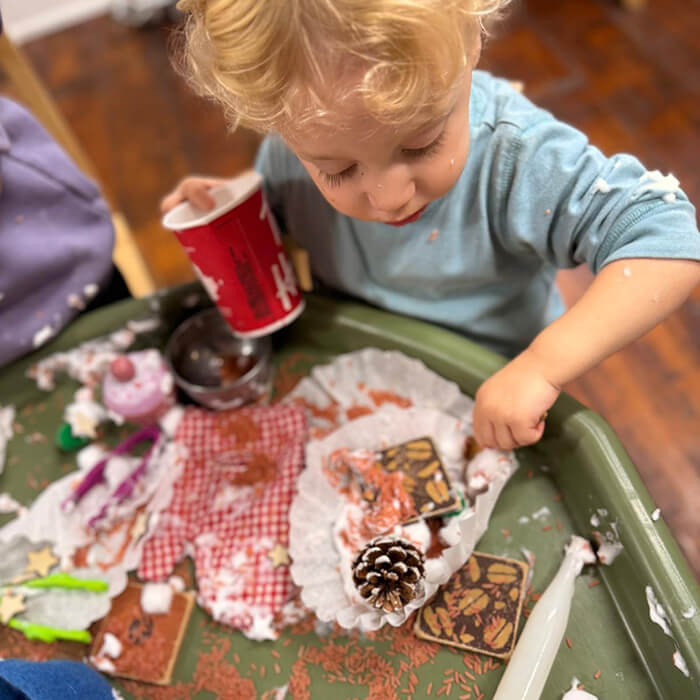 Mini Masterminds Child Care A young child plays at a table, pouring from a red cup and handling various craft items, including a pinecone and festive decorations, with some crafting mess scattered around.