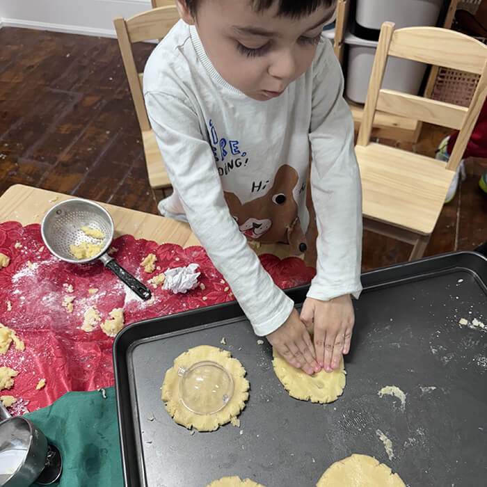 Mini Masterminds Child Care A child presses dough onto a baking tray with hands while making cookies; flour and utensils are scattered on the table.