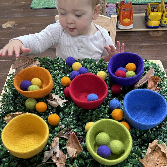 Mini Masterminds Child Care A young child plays with colorful felt balls and bowls on a table covered with green artificial leaves and dry brown leaves.