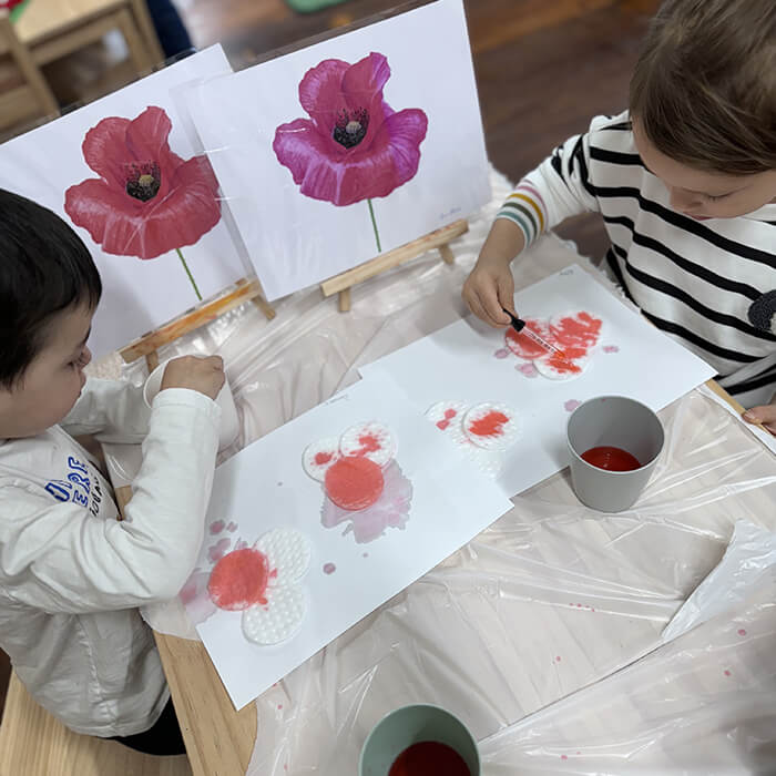 Mini Masterminds Child Care Two children sit at a table painting red flowers on paper, referencing printed images of poppies. Art supplies and cups of red paint are visible on the plastic-covered table.